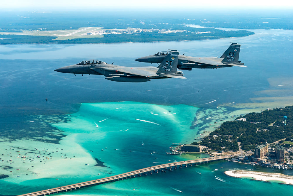 Two military aircraft flying over the blue-green water near Eglin Air Force Base.