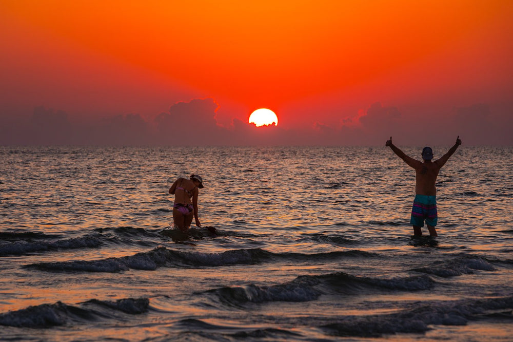Two swimmers with arms raised in the surf as the sun drops to the horizon