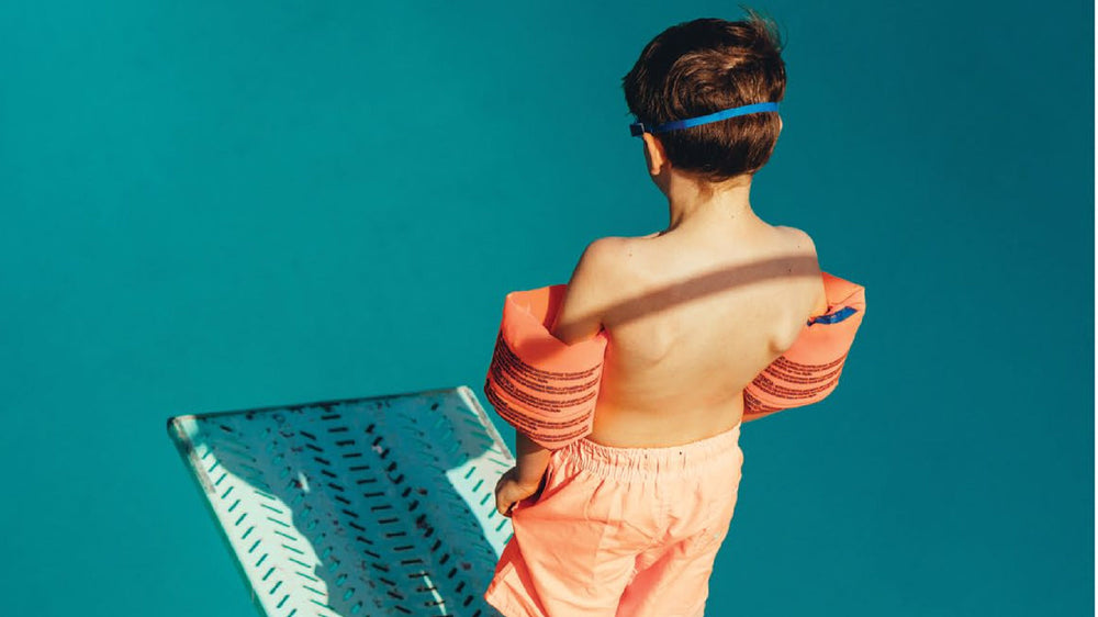 Boy in water wings standing at the end of a diving board above turquoise water