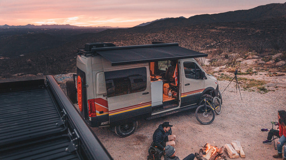 An adventure van with its awning out and friends gathered around a campfire at sunset