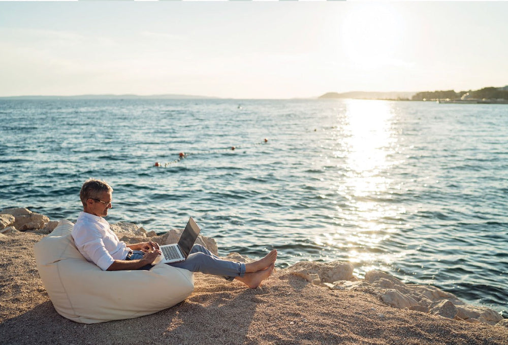 A man working on a laptop in a bean bag chair at the water's edge at sunset