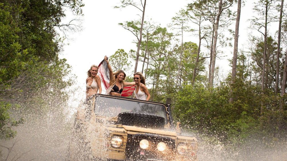 An open-top four-wheel drive blasting through a water crossing in the pines