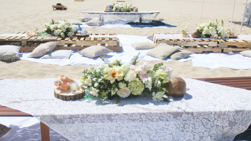A long lace-covered table set with white florals on the sand beside the water